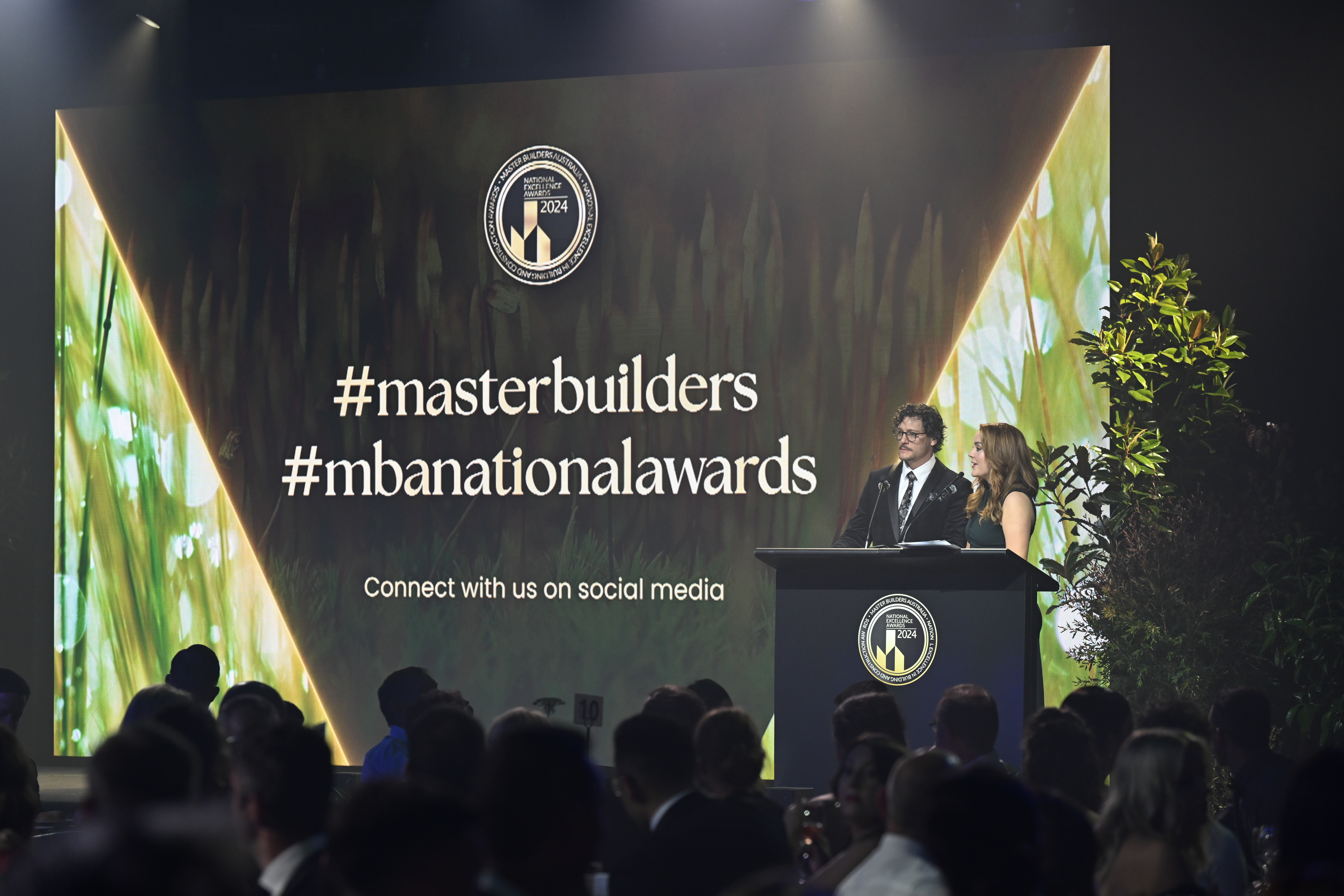 Two hosts on stage presenting at the Master Builders National Awards.